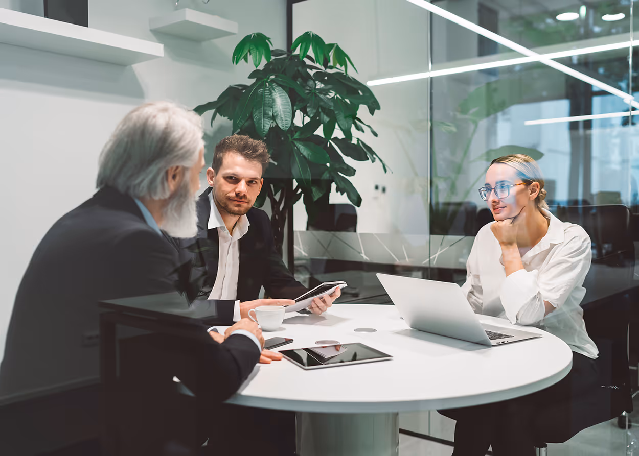 Three businesspeople having a meeting at a round white table in a modern glass office.