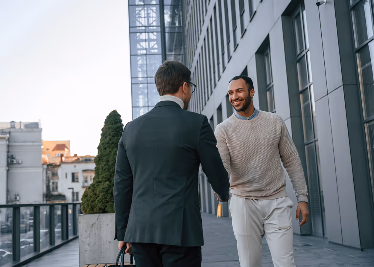 Two men shaking hands outdoors next to a modern office building, one in a suit, the other in casual clothes.