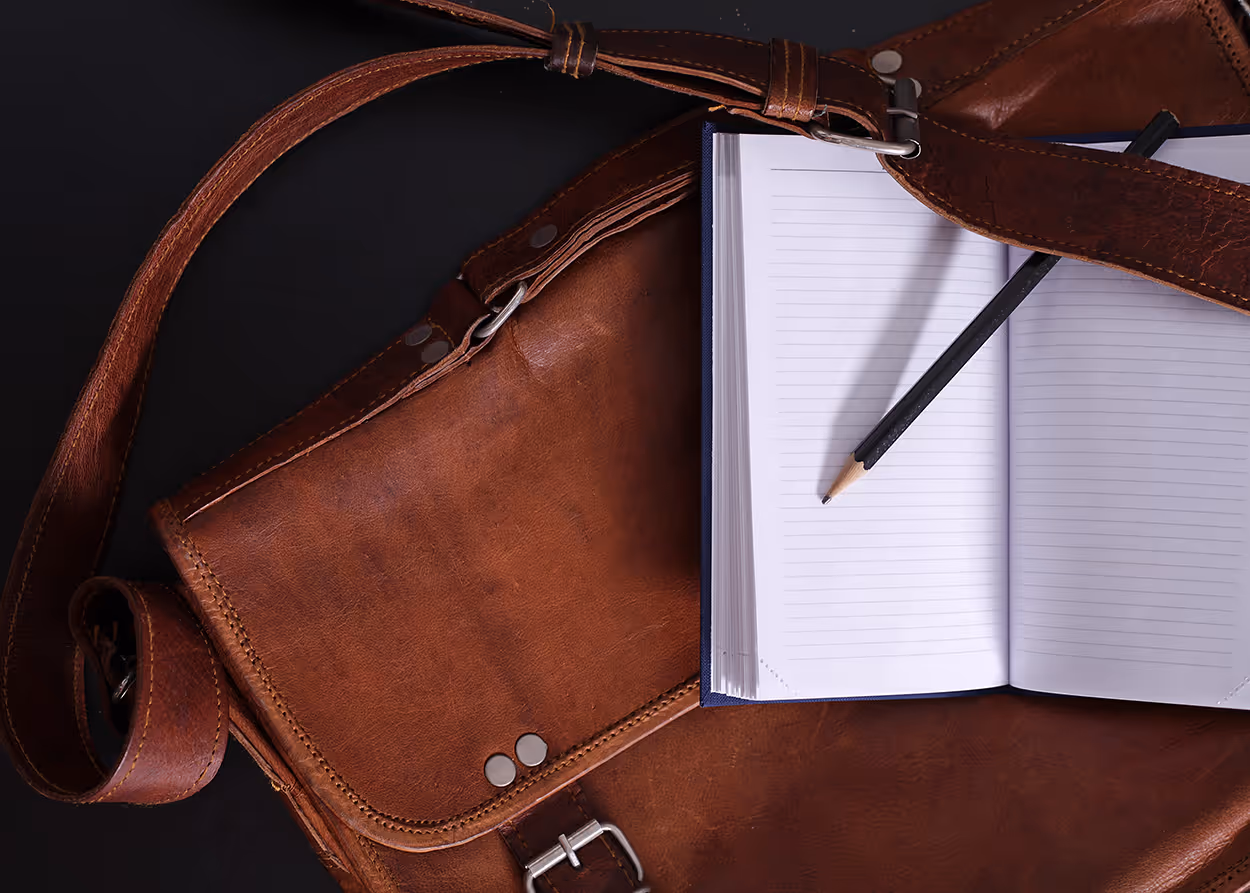 Brown leather bag with an open lined notebook and a black pencil on top, set against a black background.