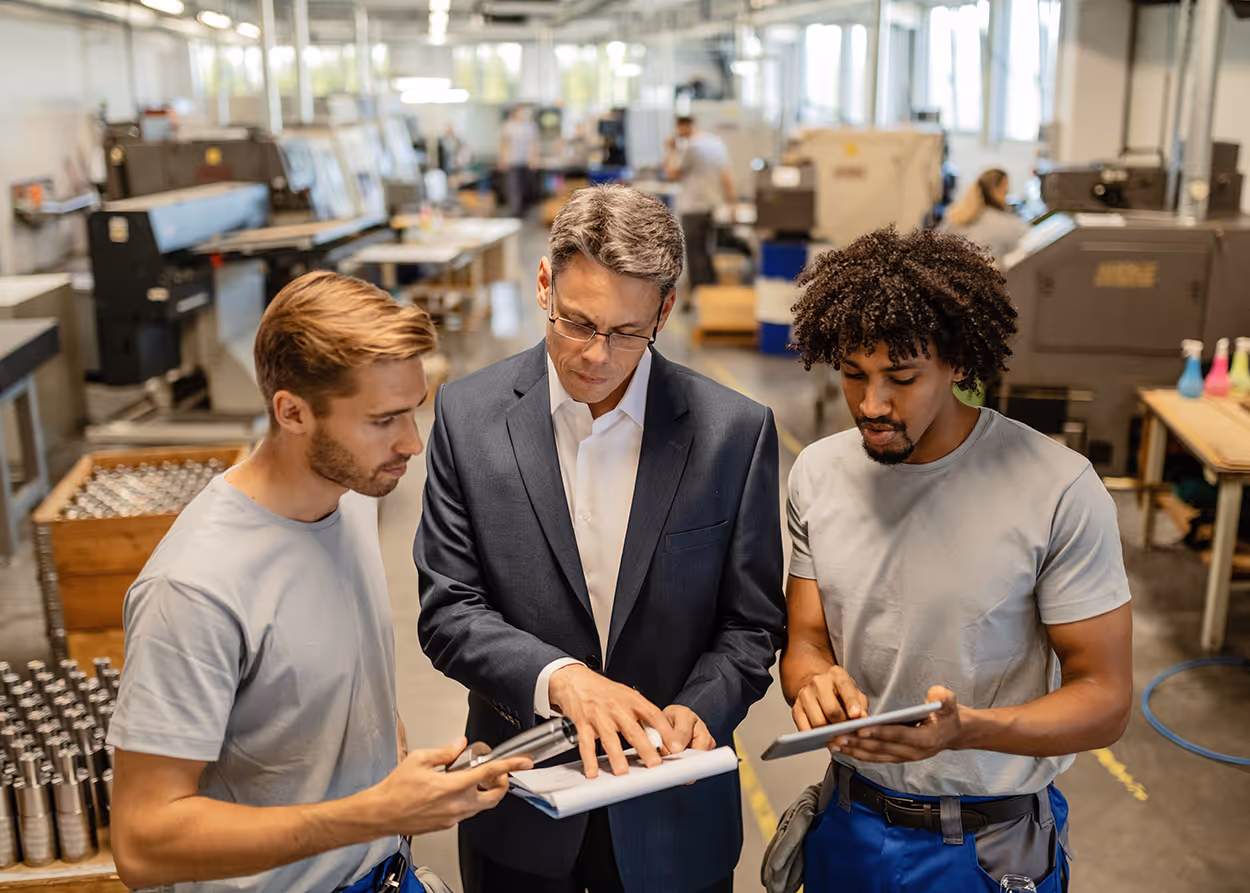 Two factory workers with tablets discussing notes with a manager in a suit inside a manufacturing facility.