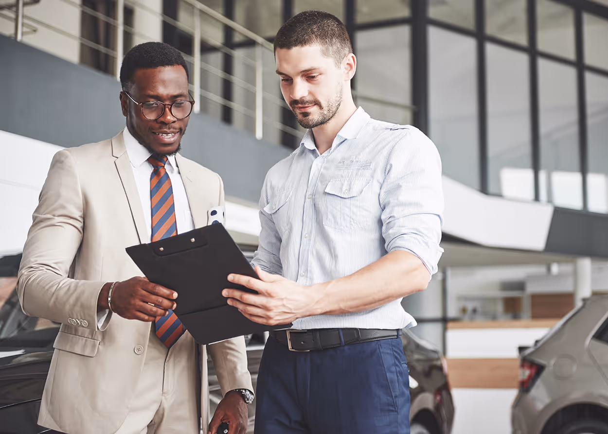 Two men discussing a document on a clipboard in a modern car showroom.
