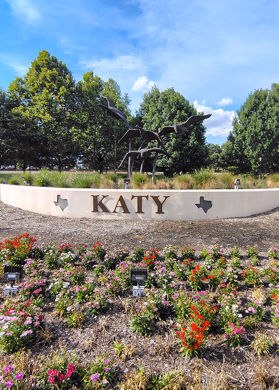 Flowerbed in front of a Katy city sign with bird sculptures and green trees under blue sky.