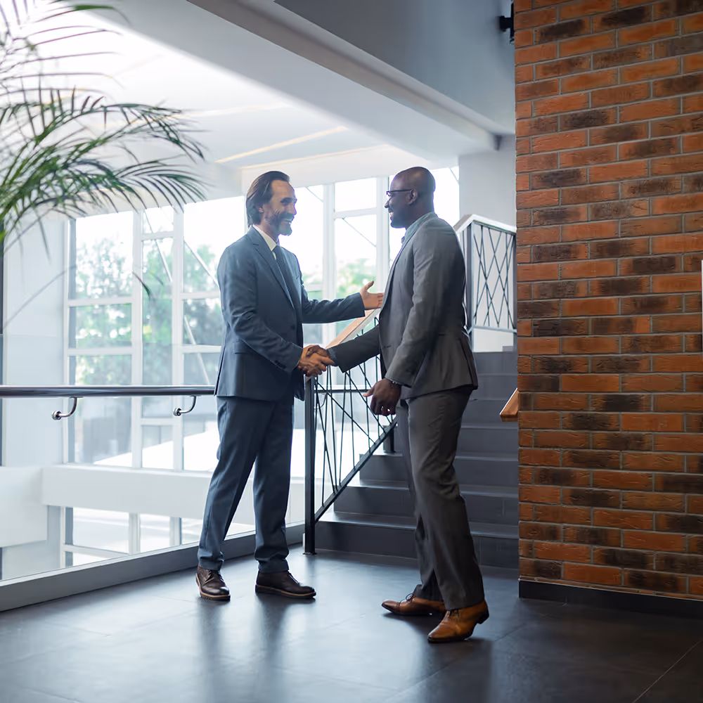 Two businessmen in suits shaking hands and smiling in a bright modern office near stairs.