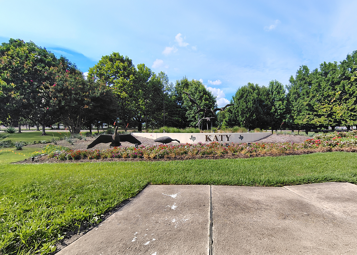 Park scene with metal bird sculptures and a sign reading 'KATY' among flower beds and green trees.