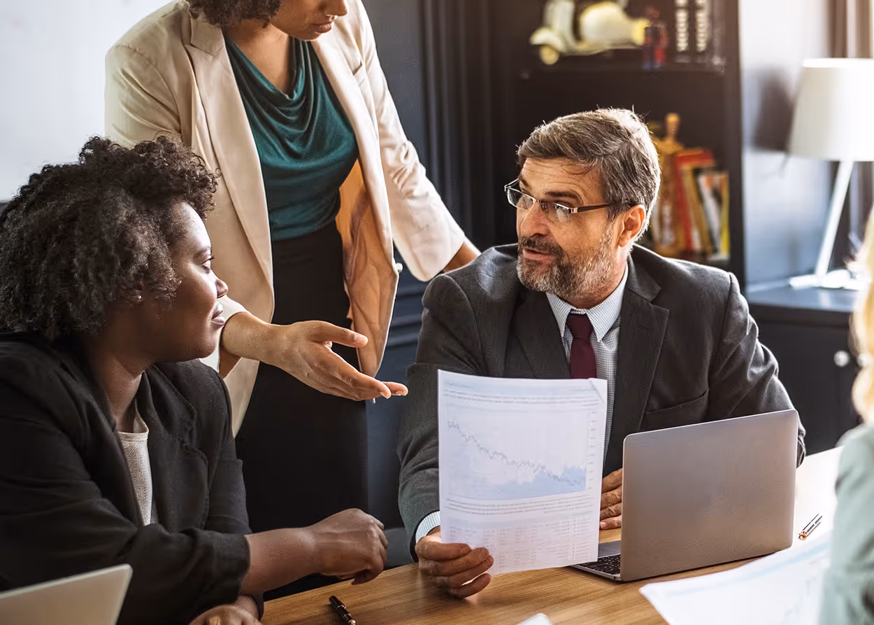 Business meeting with a man holding a chart, discussing with two women over a table with laptops.