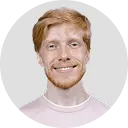 Smiling man with light red hair and beard wearing a light pink shirt against a white background.