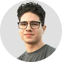 Headshot of a young man with curly hair and black glasses wearing a dark shirt, smiling slightly against a white background.