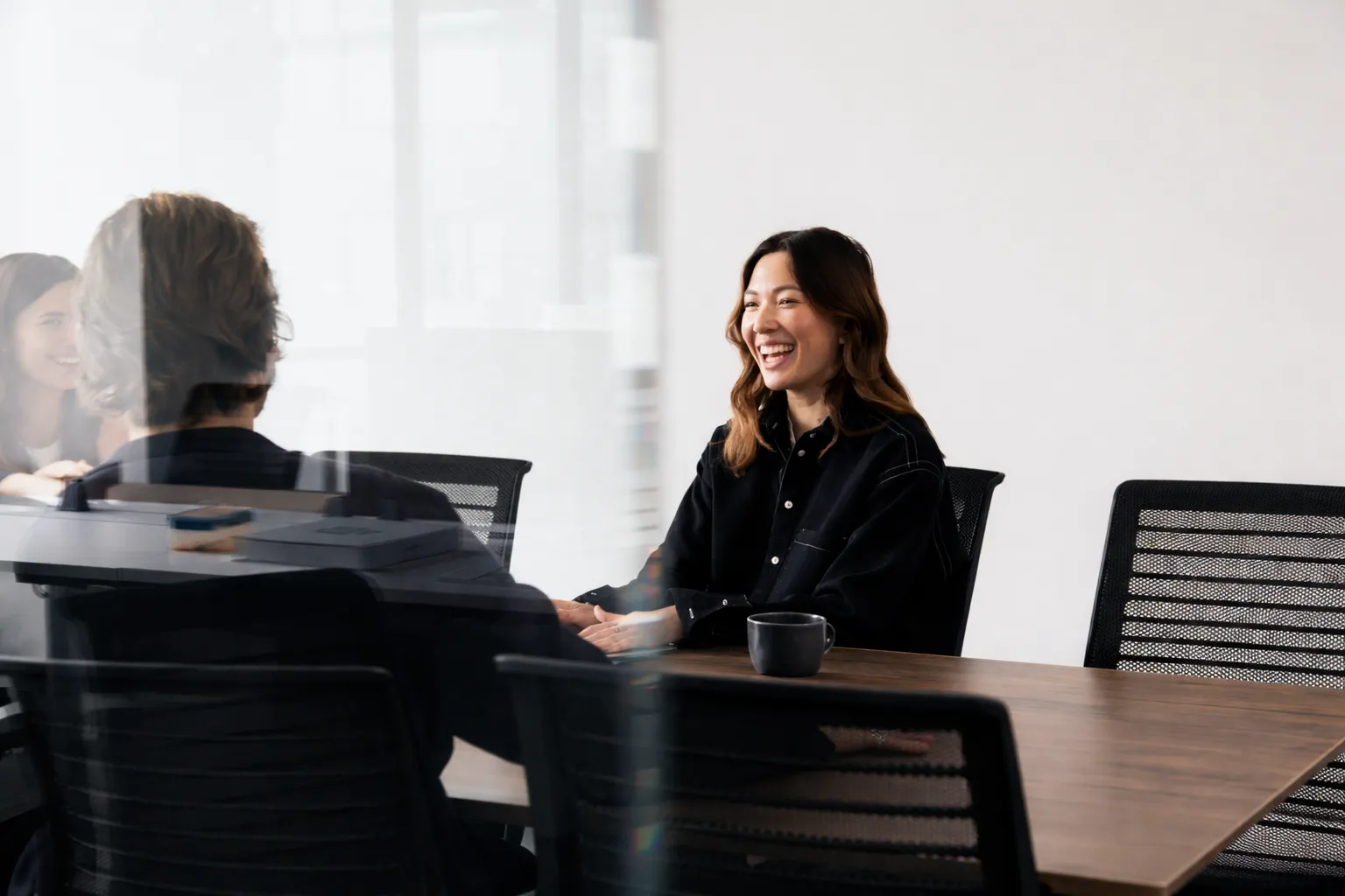 Smiling woman in a black shirt sitting at a conference table, talking with a man whose back is to the camera.