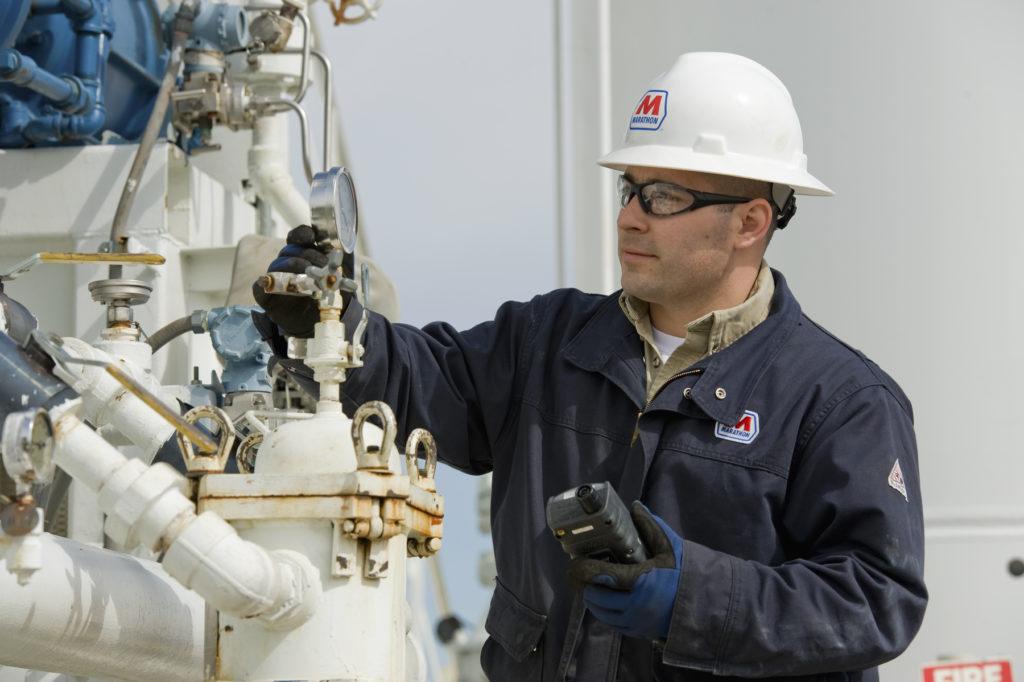 Worker in hard hat and protective glasses inspecting gauges on industrial piping equipment.