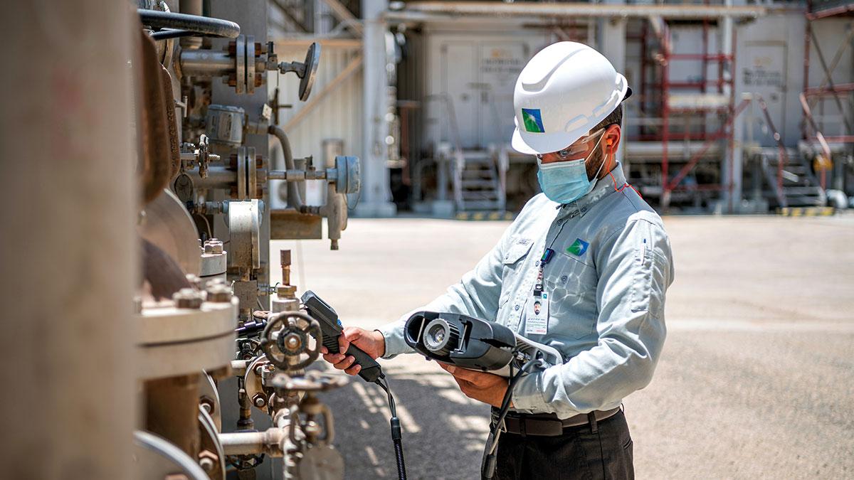 Worker in hard hat and mask inspecting industrial equipment using a handheld scanner device outdoors.