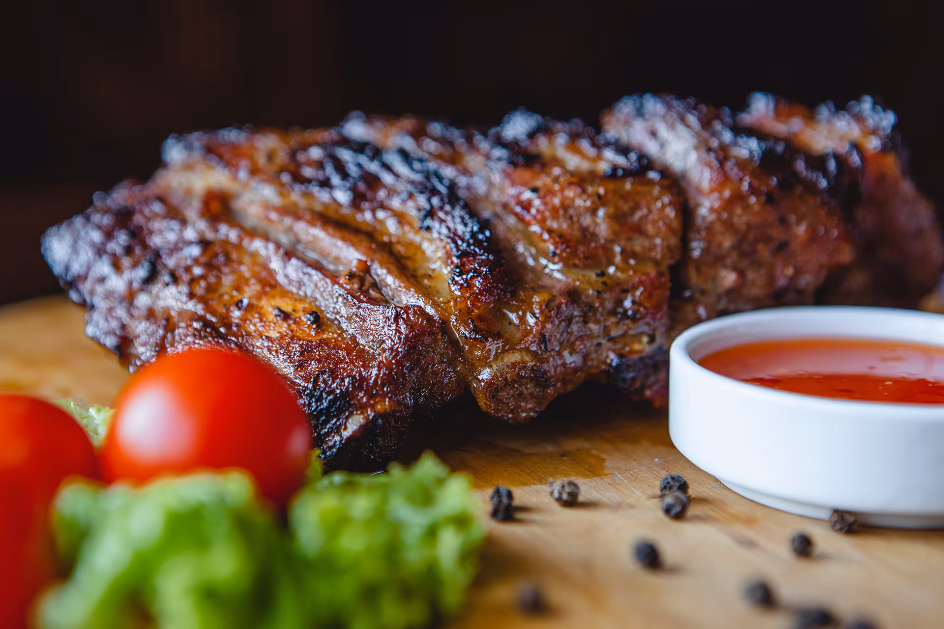 Grilled ribs with barbecue sauce, cherry tomatoes, lettuce, and black peppercorns on a wooden board.