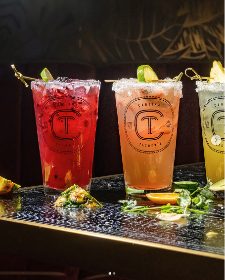 Three colorful cocktails with lime and pineapple garnishes on a dark table with herbs and fruit pieces.