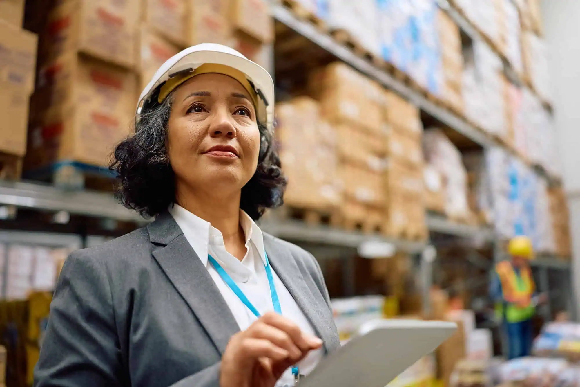 Female inspector wearing a hard hat using a tablet in a warehouse with stacked boxes.