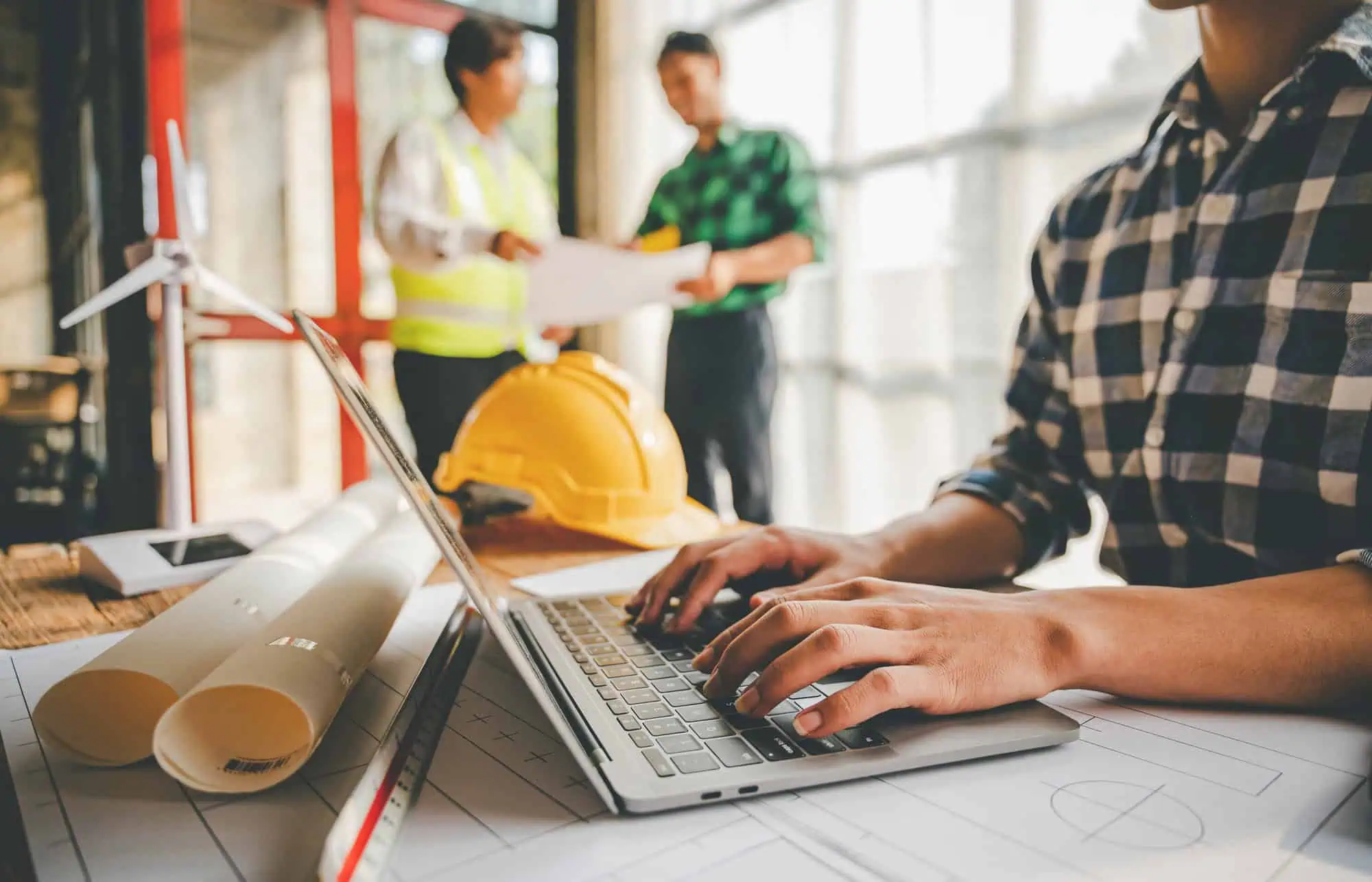 Person typing on a laptop with blueprints, a hard hat, and a scale ruler on the table and two people discussing plans.