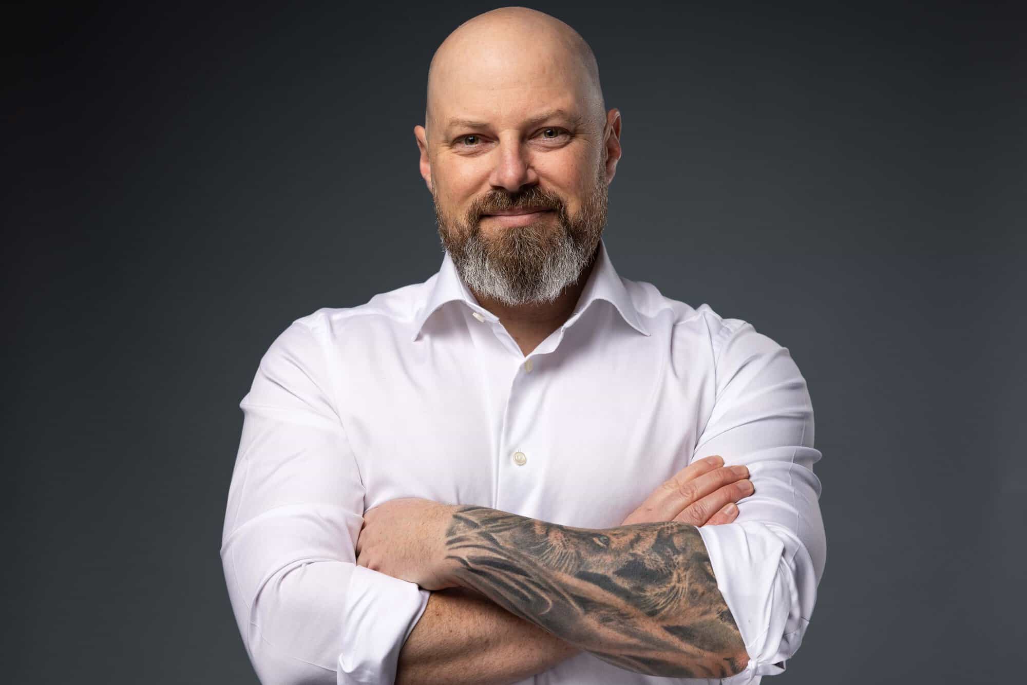Bald man with a beard wearing a white shirt, arms crossed showing a lion tattoo on his forearm.