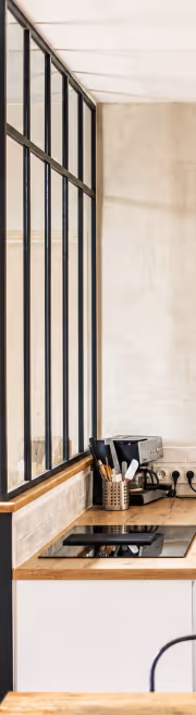 Coin de cuisine minimaliste avec plan de travail en bois, plaque de cuisson induite, machine à café et accessoires près d'une grande fenêtre à cadres métalliques.