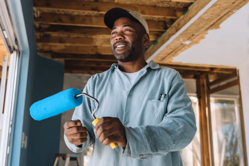 Smiling painter holding a blue paint roller inside a room under renovation.
