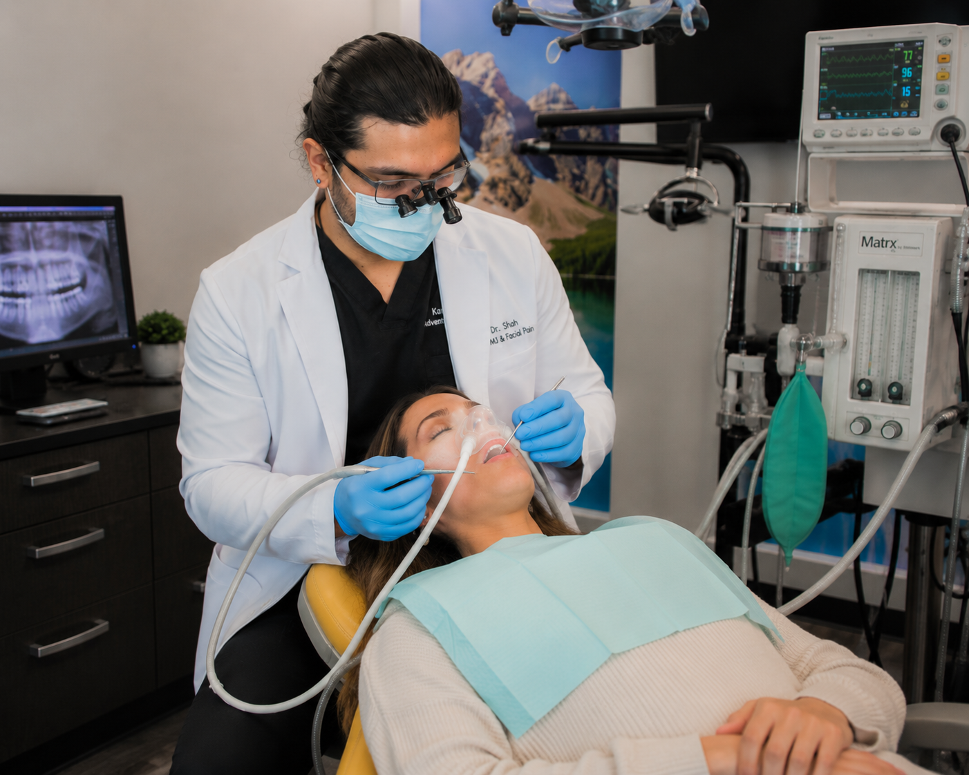 Dr. Karnik Shah performing a dental procedure on a sedated female patient wearing a nitrous oxide mask with a vital signs monitor in the background at Comfydent Smiles in Bee Cave, TX.
