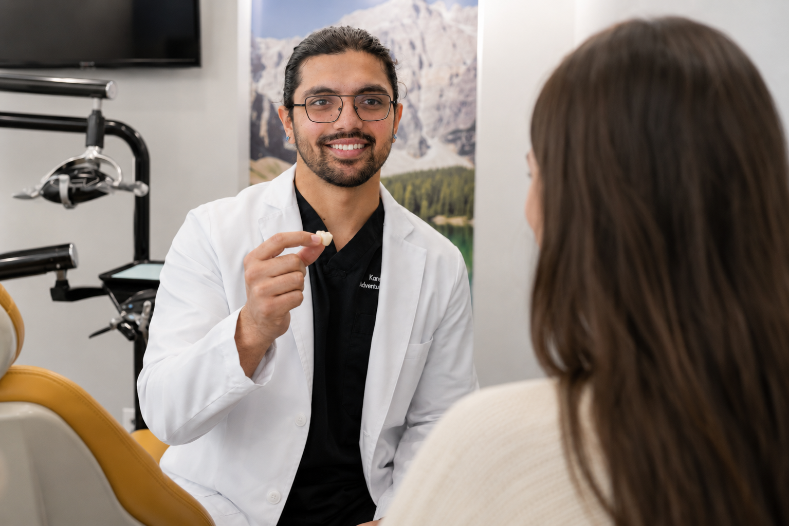 Dr. Karnik Shah at ComfyDent Smiles in Bee Cave, TX, smiling and holding a dental crown sample while explaining treatment options to a patient in the exam chair.