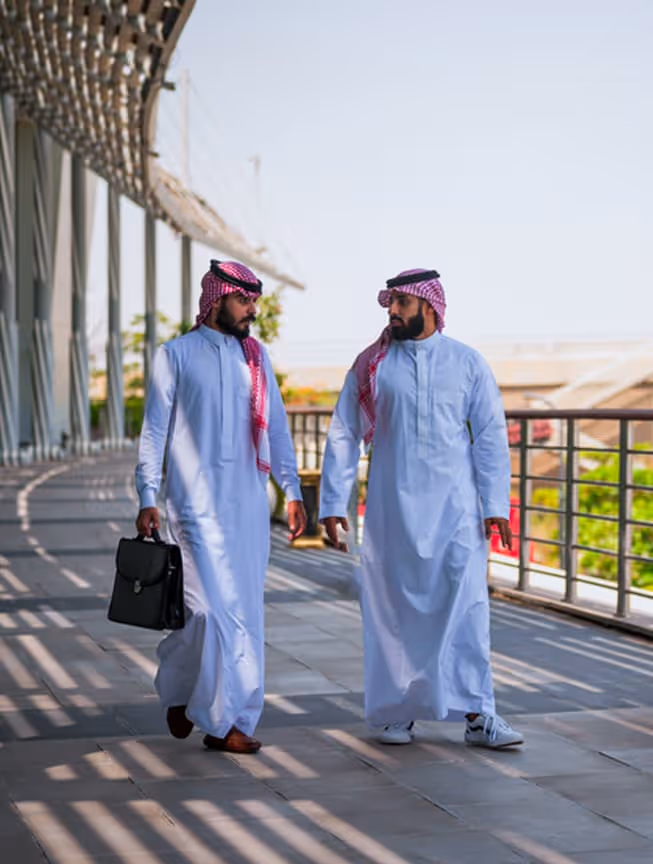 رجلان سعوديان يمشيان ويتحدثان في ممر مفتوح بأزياء تقليدية ويحملان حقيبة ويدخلان الظل.