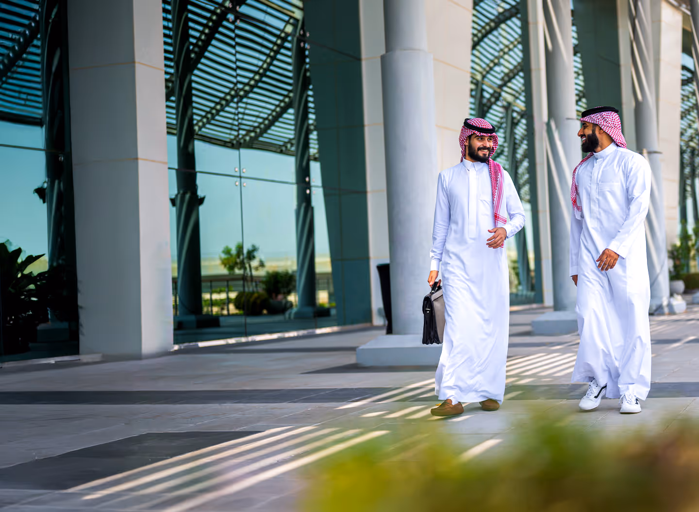 رجلان سعوديان يرتديان الثوب والغترة يتحدثان أثناء المشي في مبنى حديث بزجاج وأعمدة.
