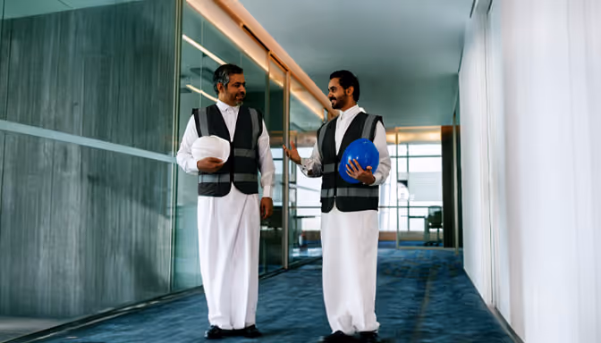 رجلان يرتديان ملابس سعودية ويحملان خوذات أمان ويتحدثان في ممر مكتب حديث.