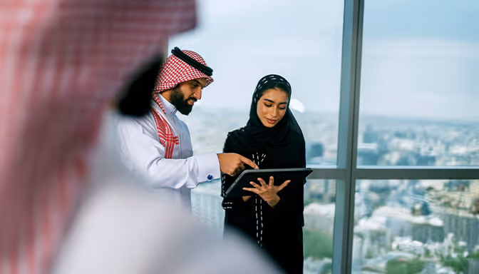 رجل وامرأة سعوديان يناقشان شيئًا على جهاز لوحي في مكتب مقابل نافذة تطل على المدينة.