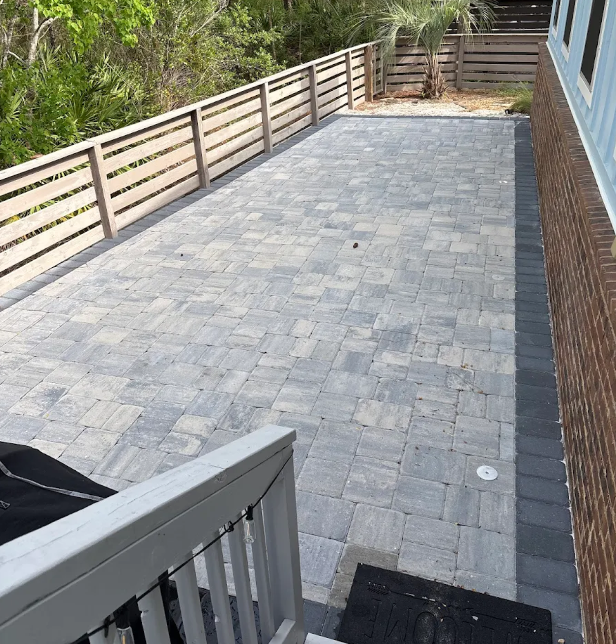 Gray stone patio beside blue house with wooden fence and green foliage in background.