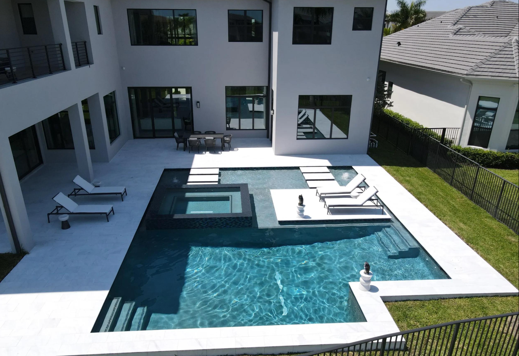 Modern backyard pool with built-in hot tub, lounge chairs, and white tiled patio beside a house.