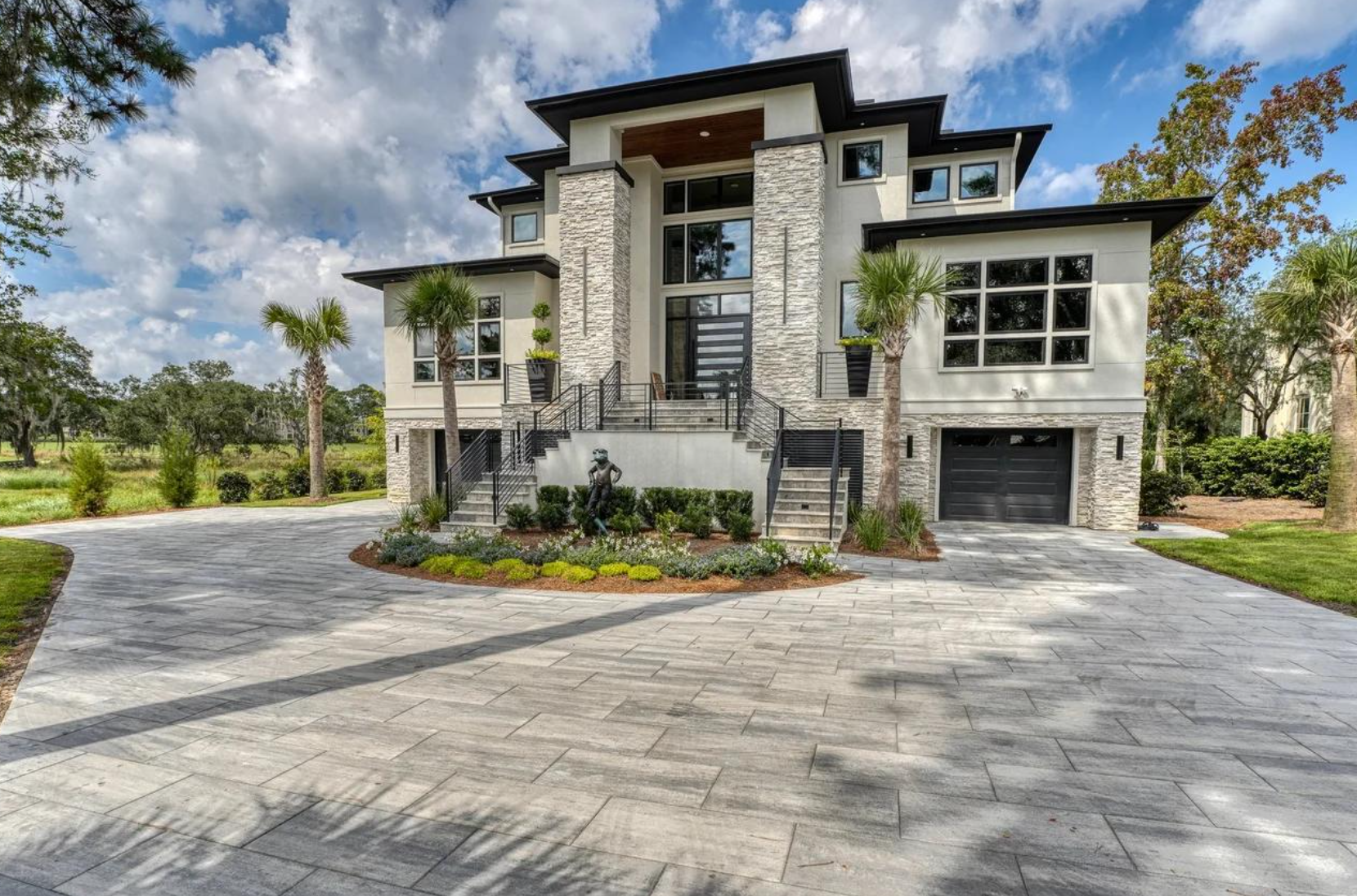 Modern two-story house with stone facade, large driveway, palm trees, and landscaped garden.