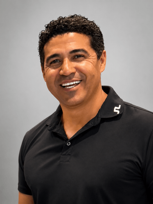 Smiling man with curly hair wearing a black polo shirt against a gray background.