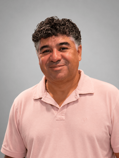 Man with curly black and gray hair wearing a light pink polo shirt and smiling at the camera.