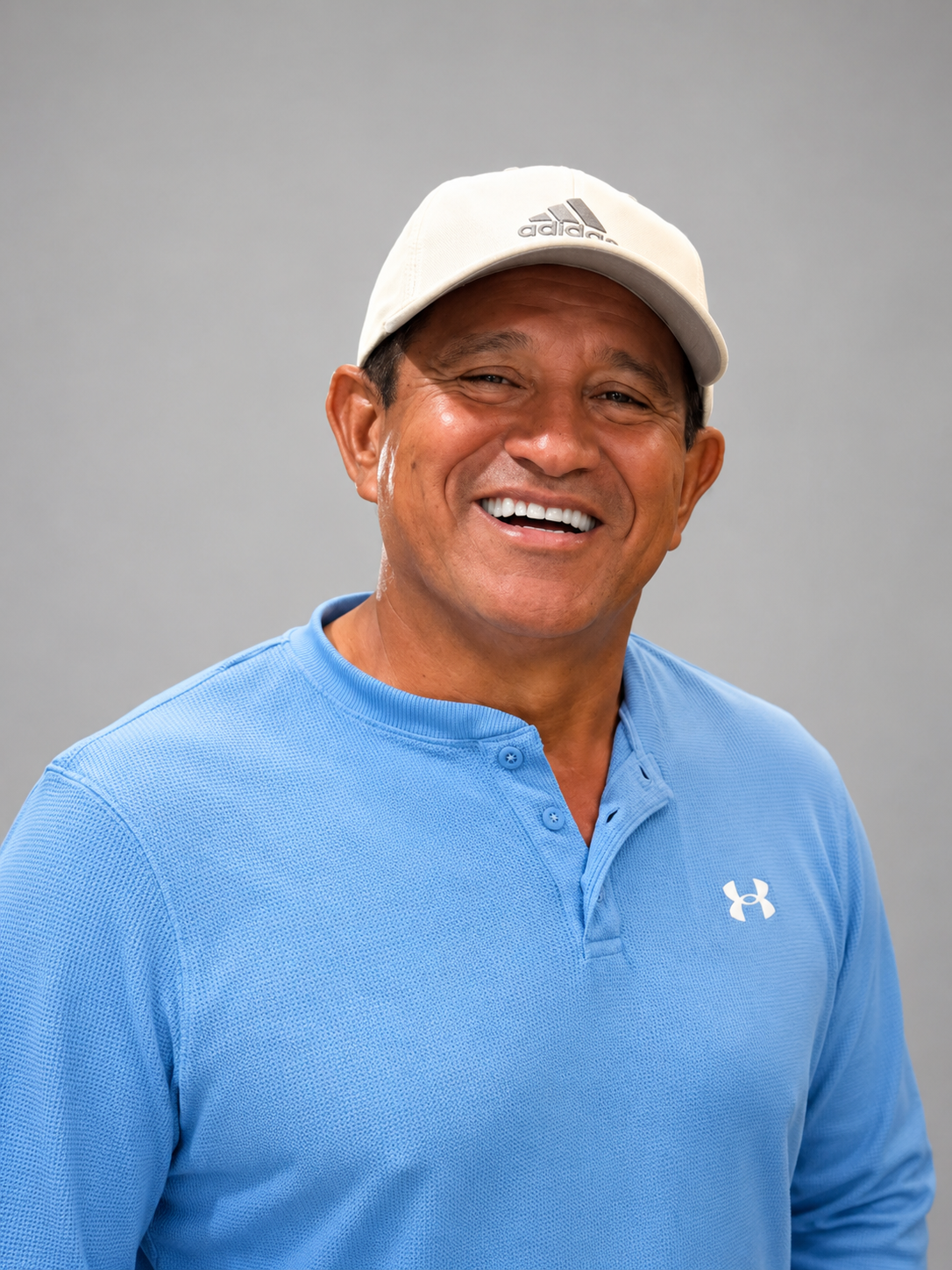 Smiling man wearing a white Adidas cap and blue Under Armour shirt against a gray background.