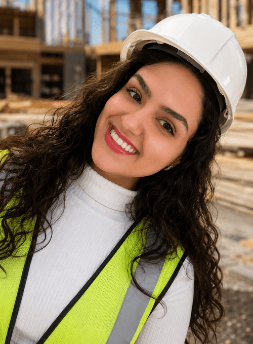 Smiling woman wearing a white hard hat and yellow safety vest at a construction site.