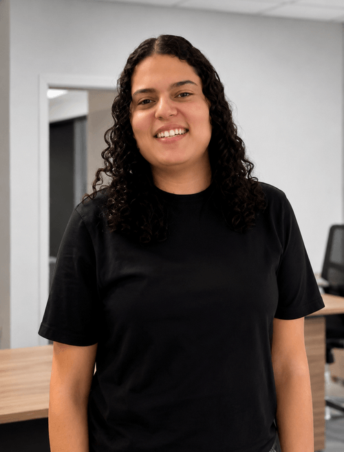 Smiling person with curly dark hair wearing a black t-shirt standing in an office setting.