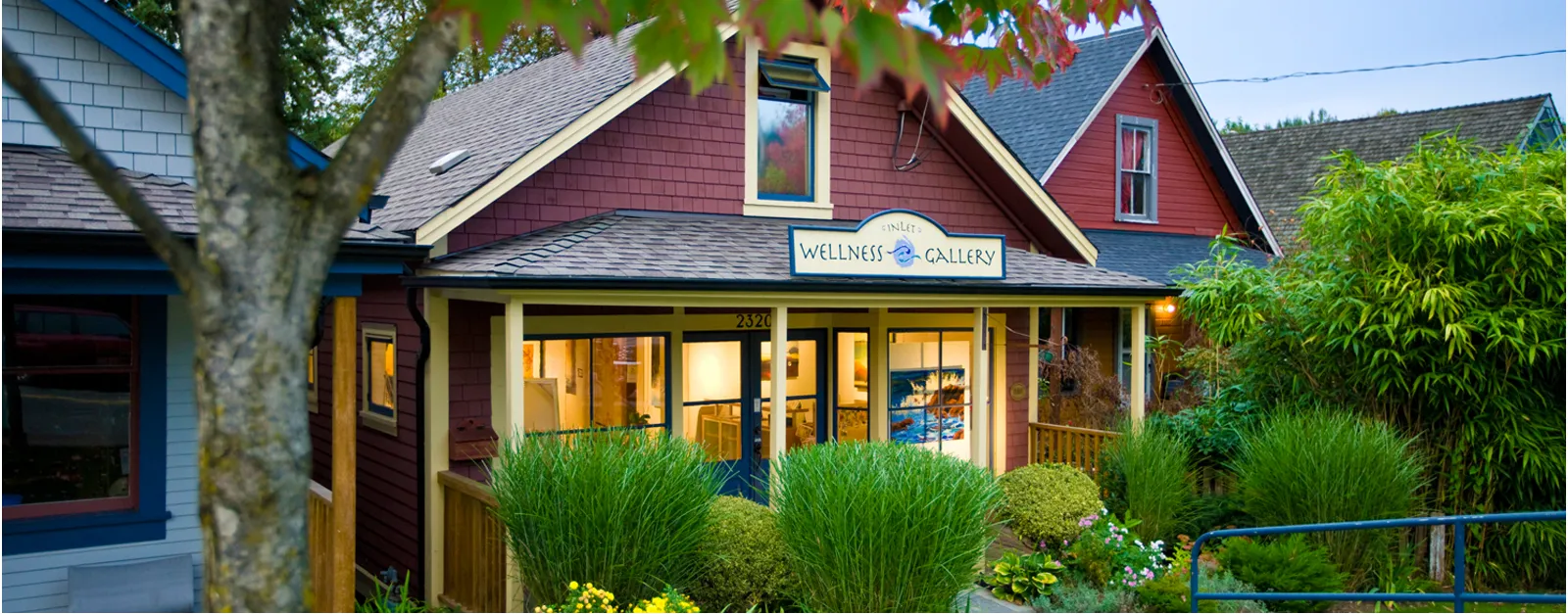 Exterior of a small Wellness Gallery, Inlet Hearing, with warm interior lights and lush green landscaping in front.