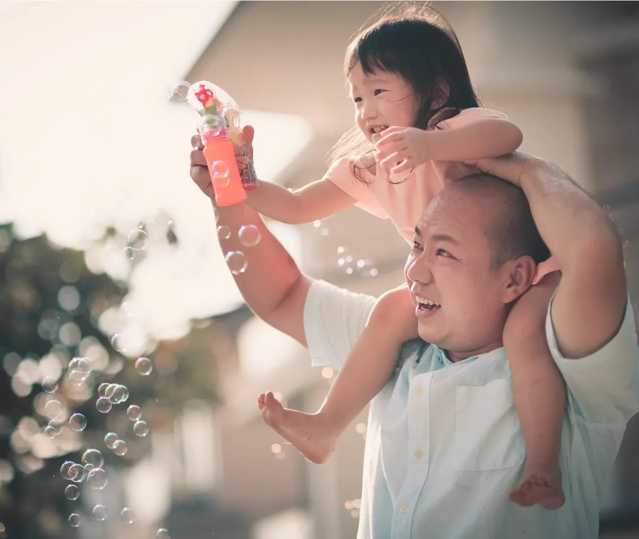A man carrying a smiling girl on his shoulders as she blows bubbles outdoors.