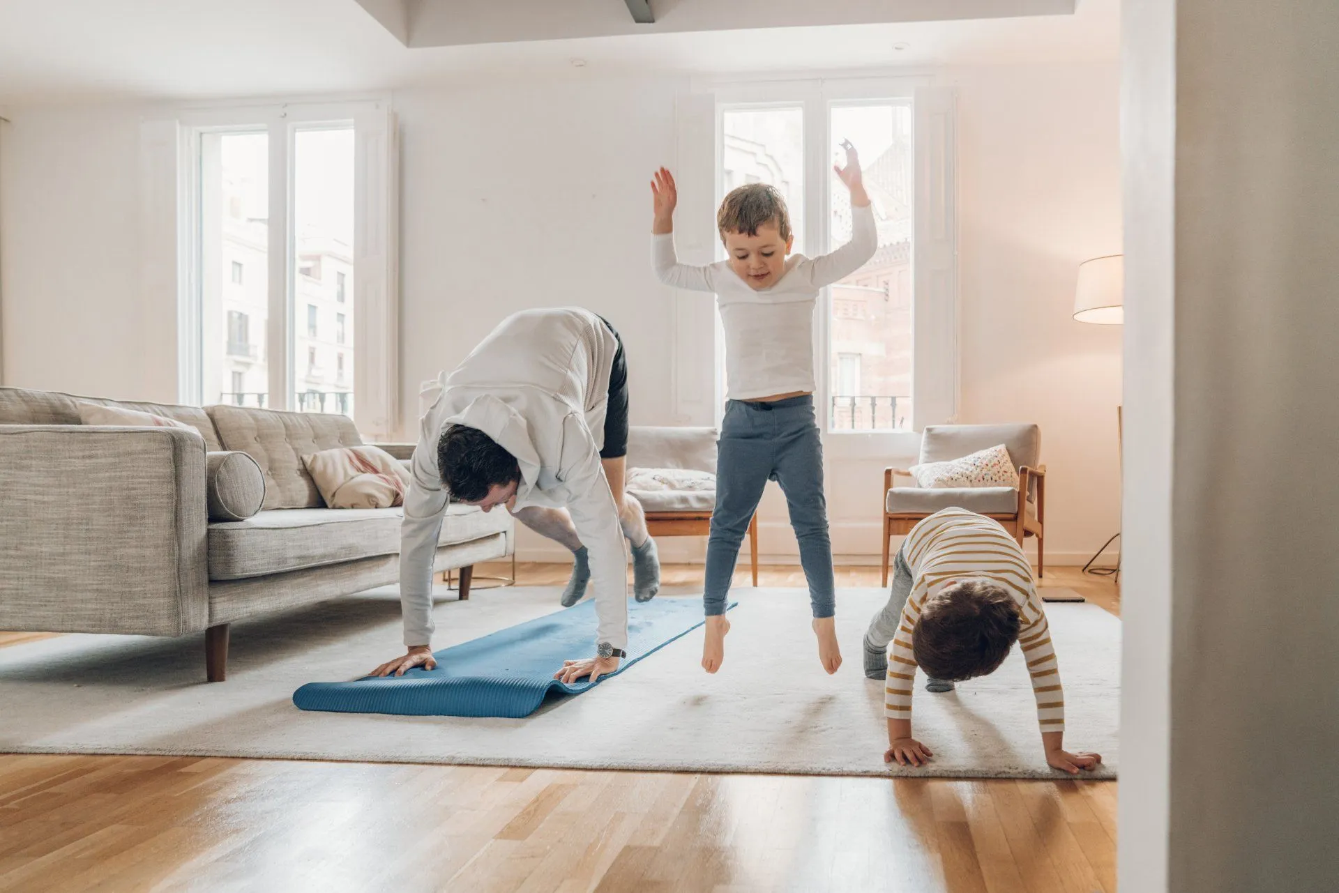 Adult and two children exercising on a rug in a bright living room with large windows.