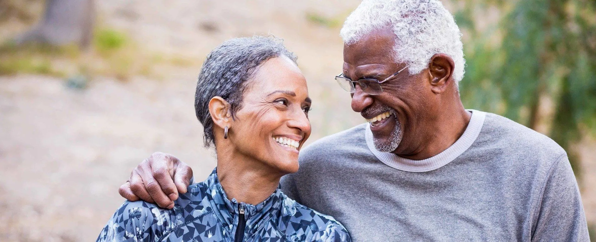 Smiling older couple looking at each other outdoors, man with arm around woman's shoulder.