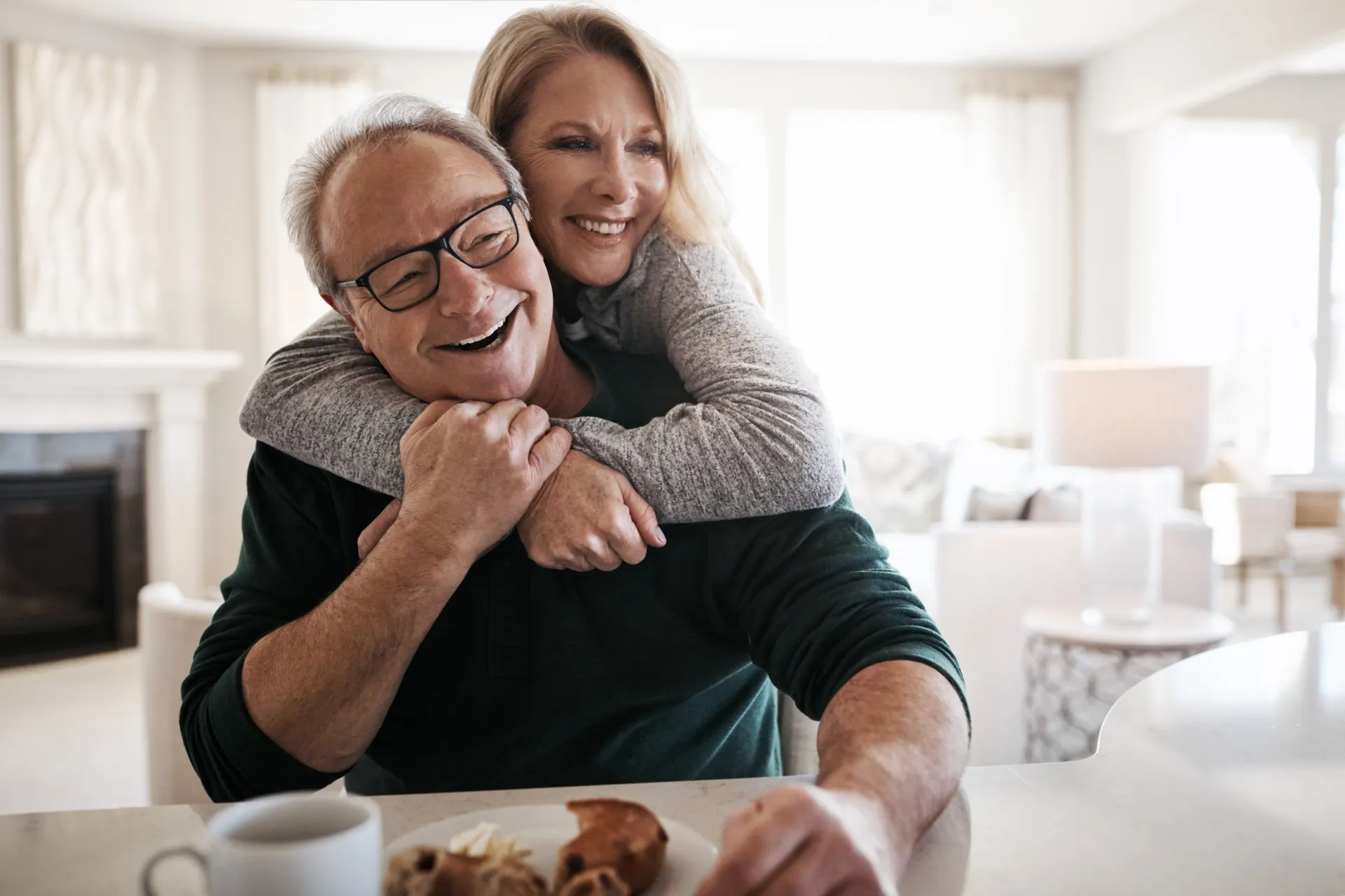 Smiling middle-aged woman hugs man wearing glasses from behind in a bright kitchen.