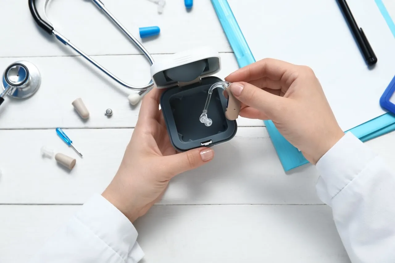 Hands holding a hearing aid and its storage case on a white medical desk with stethoscope and tools.