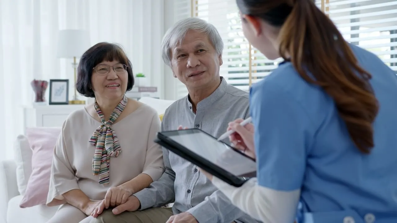 Elderly couple holding hands while talking to a nurse in a bright living room.