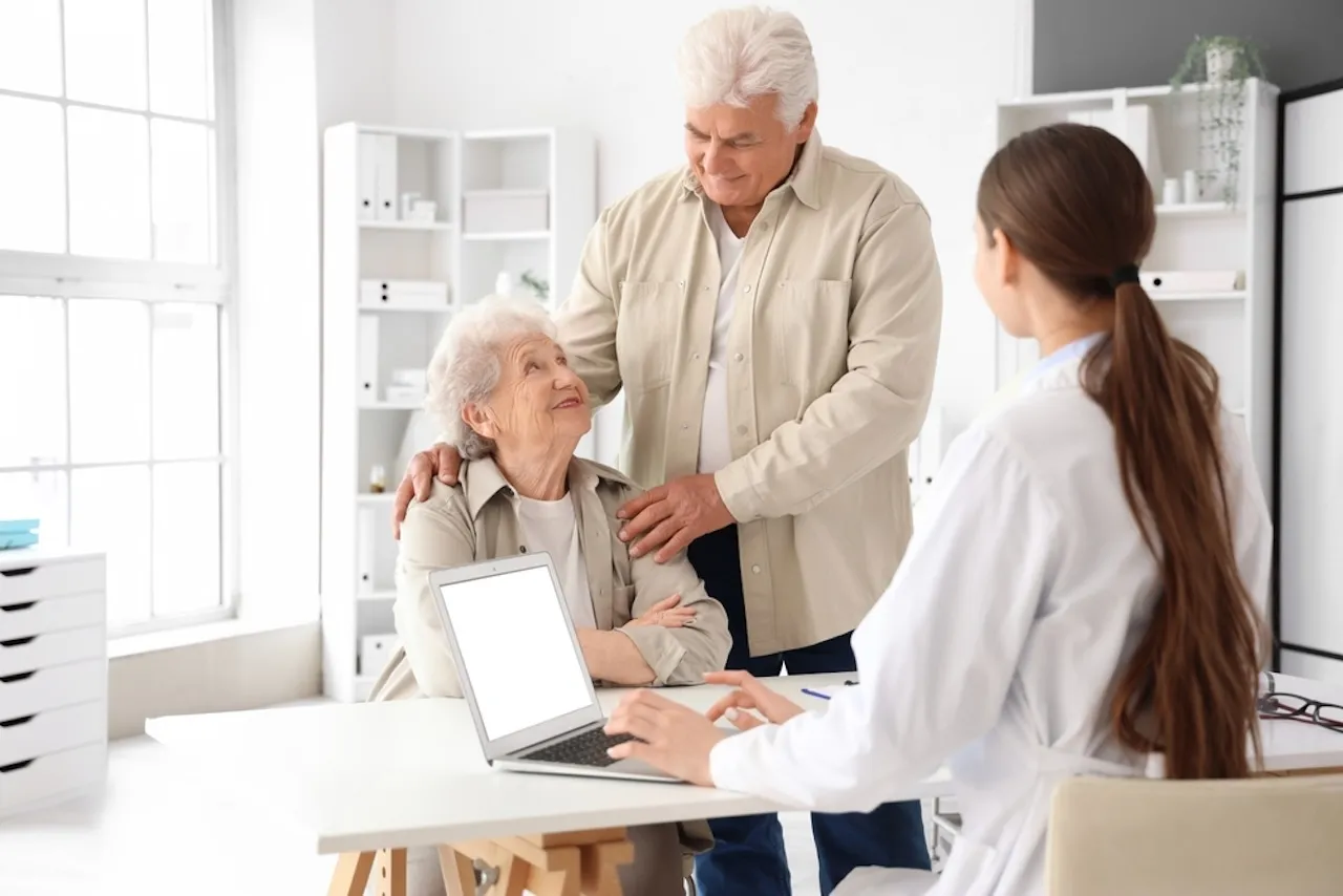 Elderly woman and man smiling at each other while a female doctor types on laptop in a bright office.
