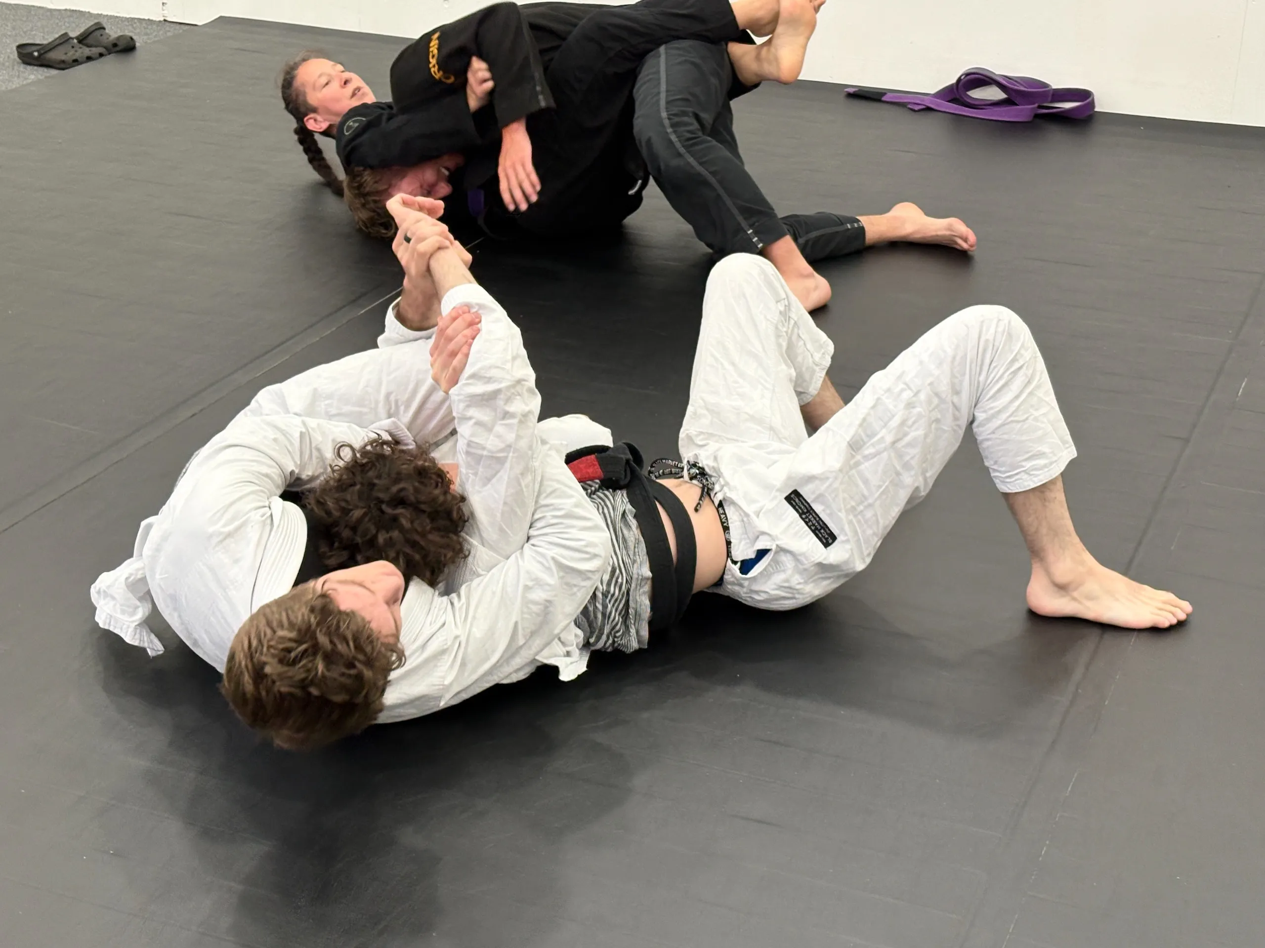 Two pairs of martial artists spar on black mats, wearing gis with one pair grappling on the floor.