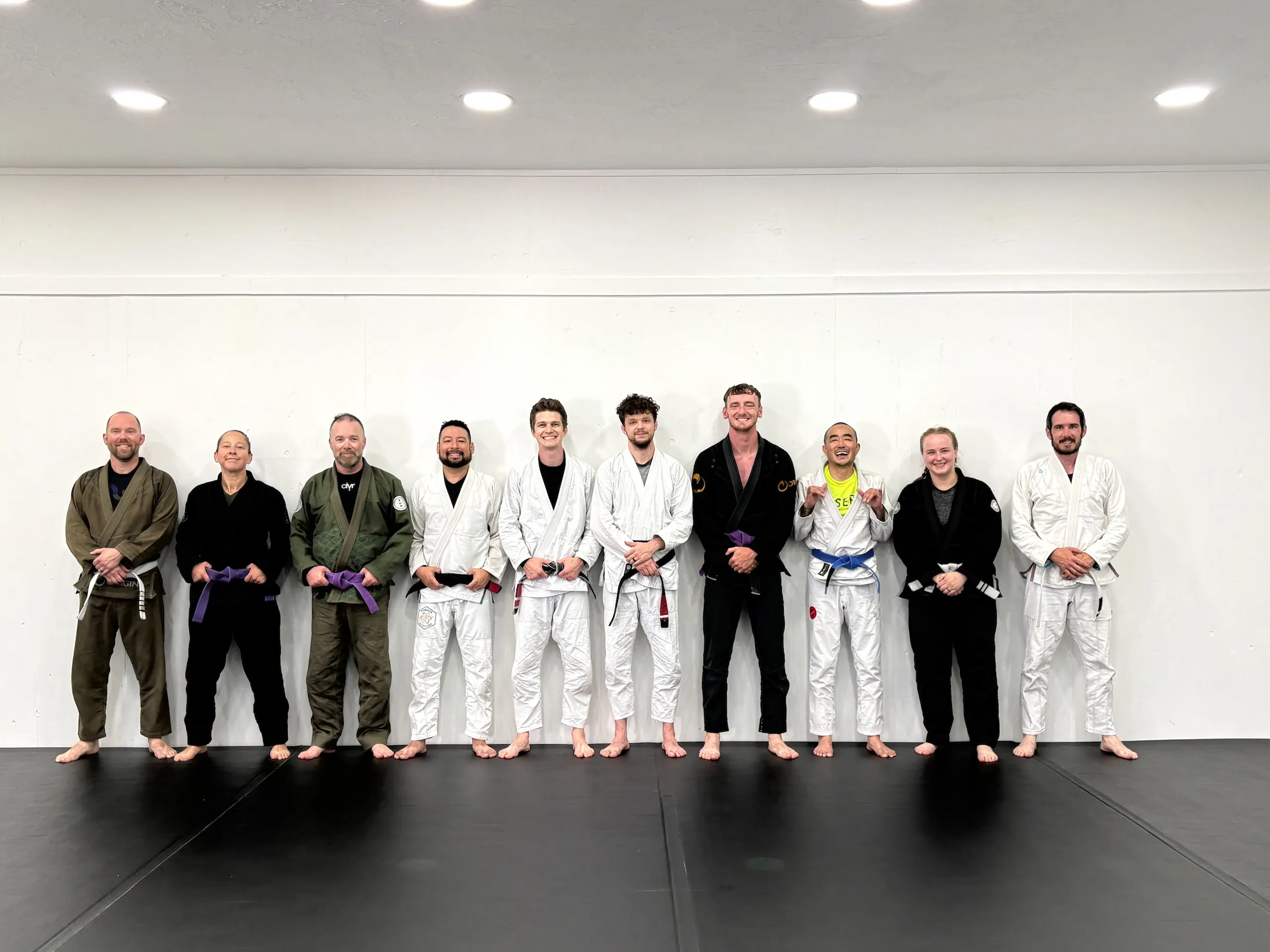 Group of nine martial artists standing barefoot in gis with different colored belts on mats.