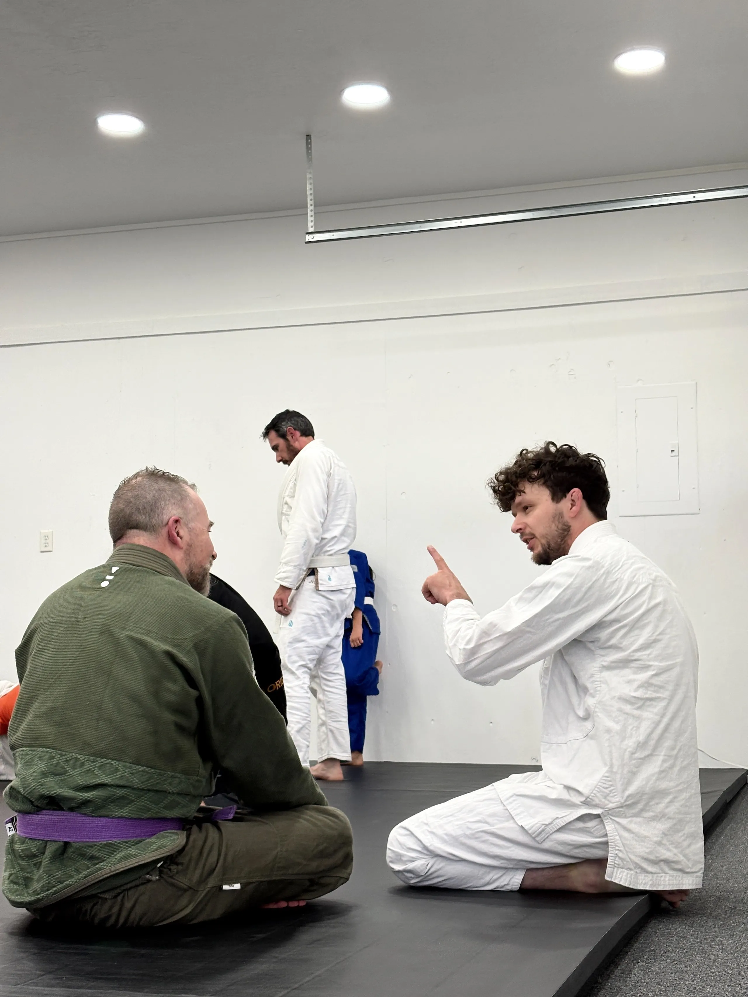 Two men in martial arts gis sit on mats talking; two others stand in the background in gis.
