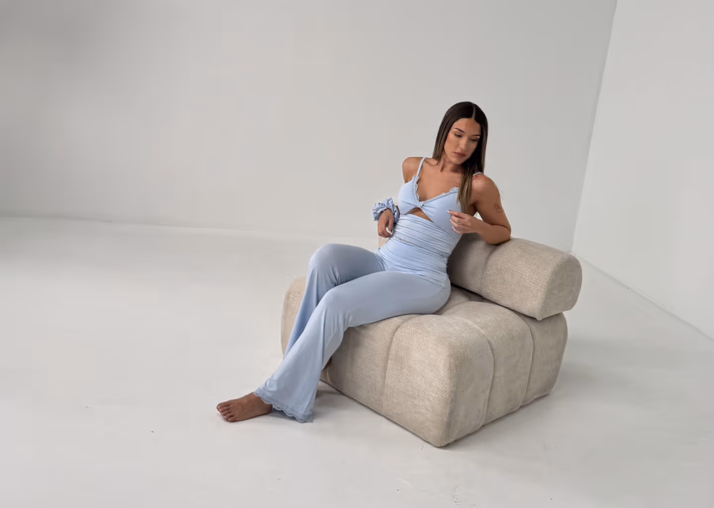 Woman in light blue outfit sitting barefoot on a textured beige lounge chair in a minimalist room.