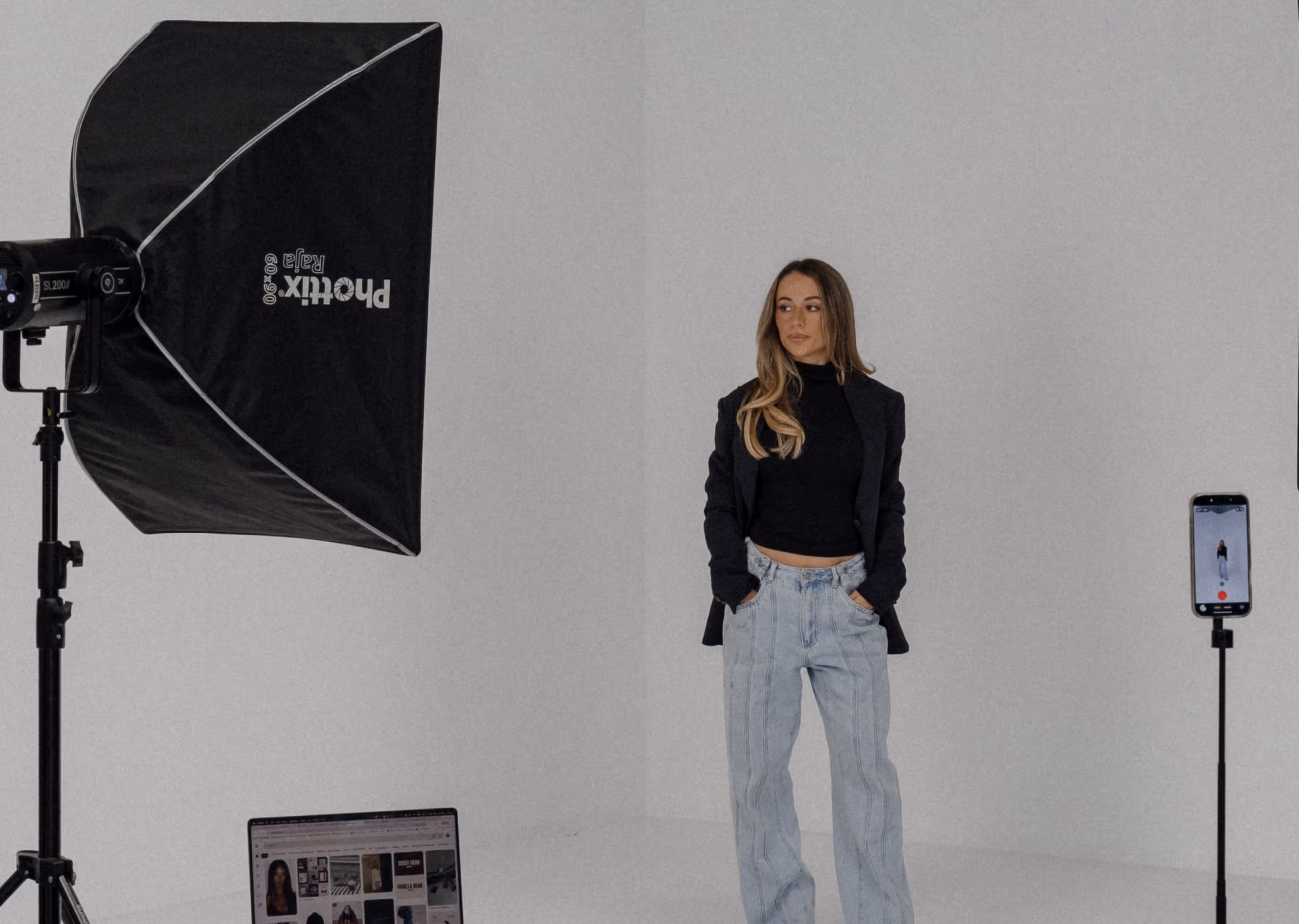 Woman in black top, blazer, and jeans posing in a studio with professional lighting and phone camera setup.