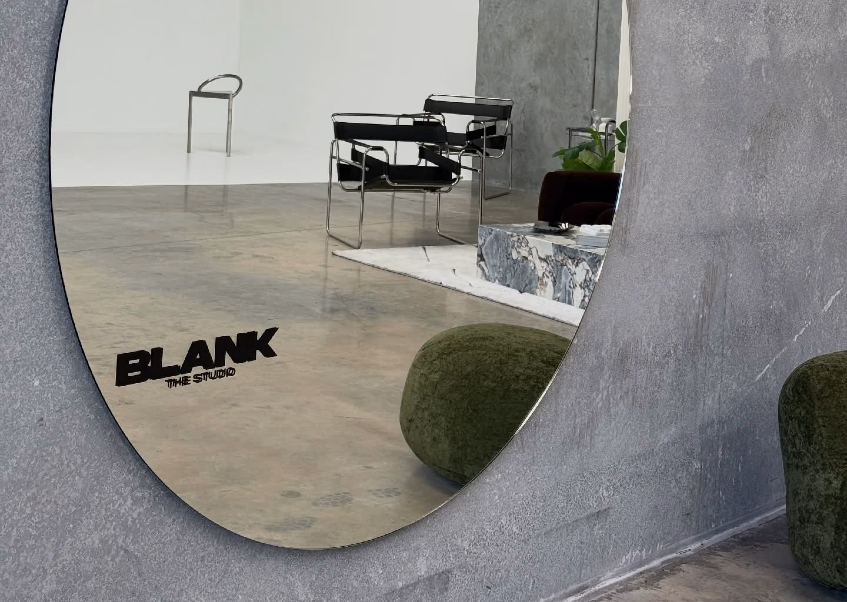 Oval mirror reflecting modern black chairs, a marble table, and a green velvet pouf in a minimalist room.