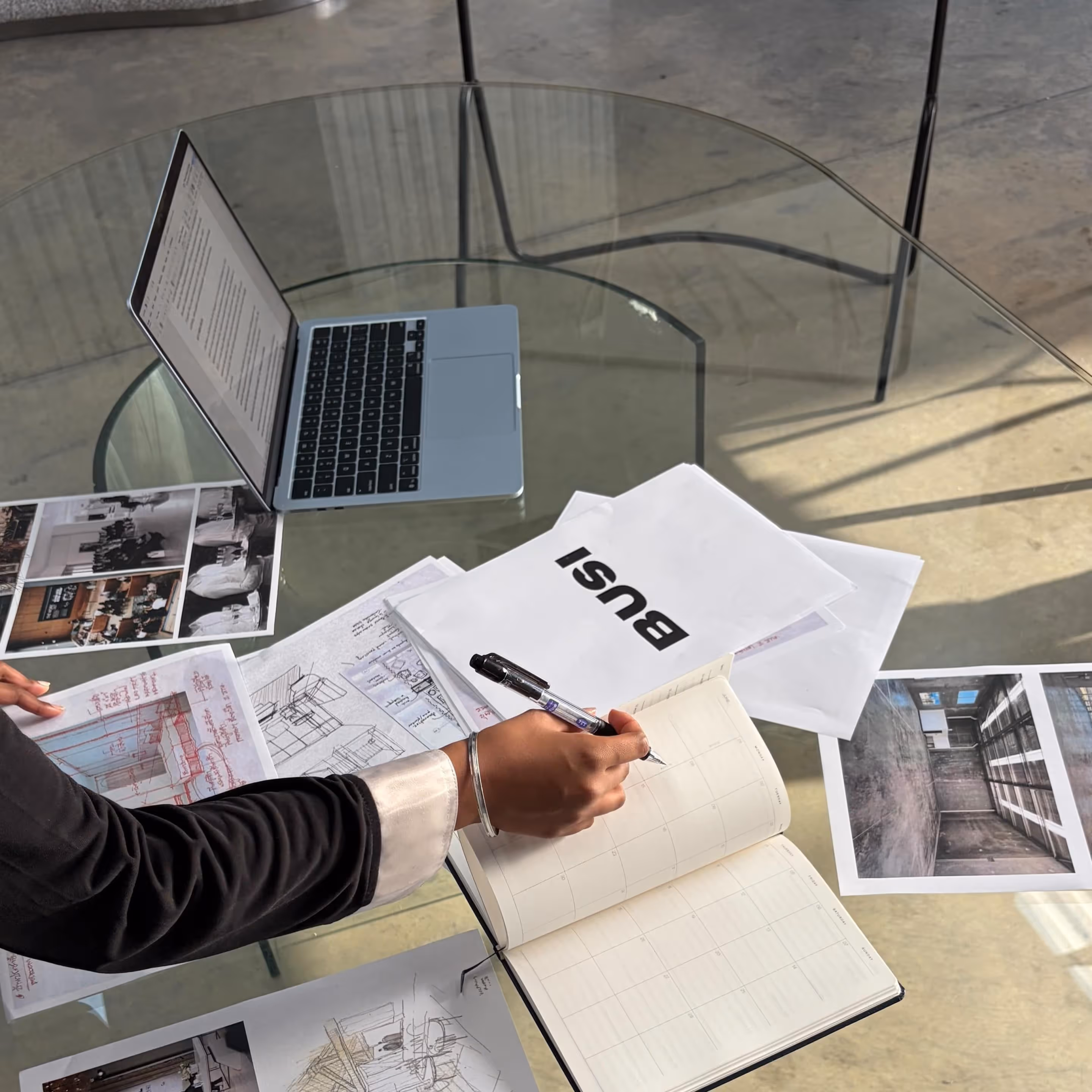 Person writing in an open planner at a glass table with architectural sketches, photos, and a laptop.